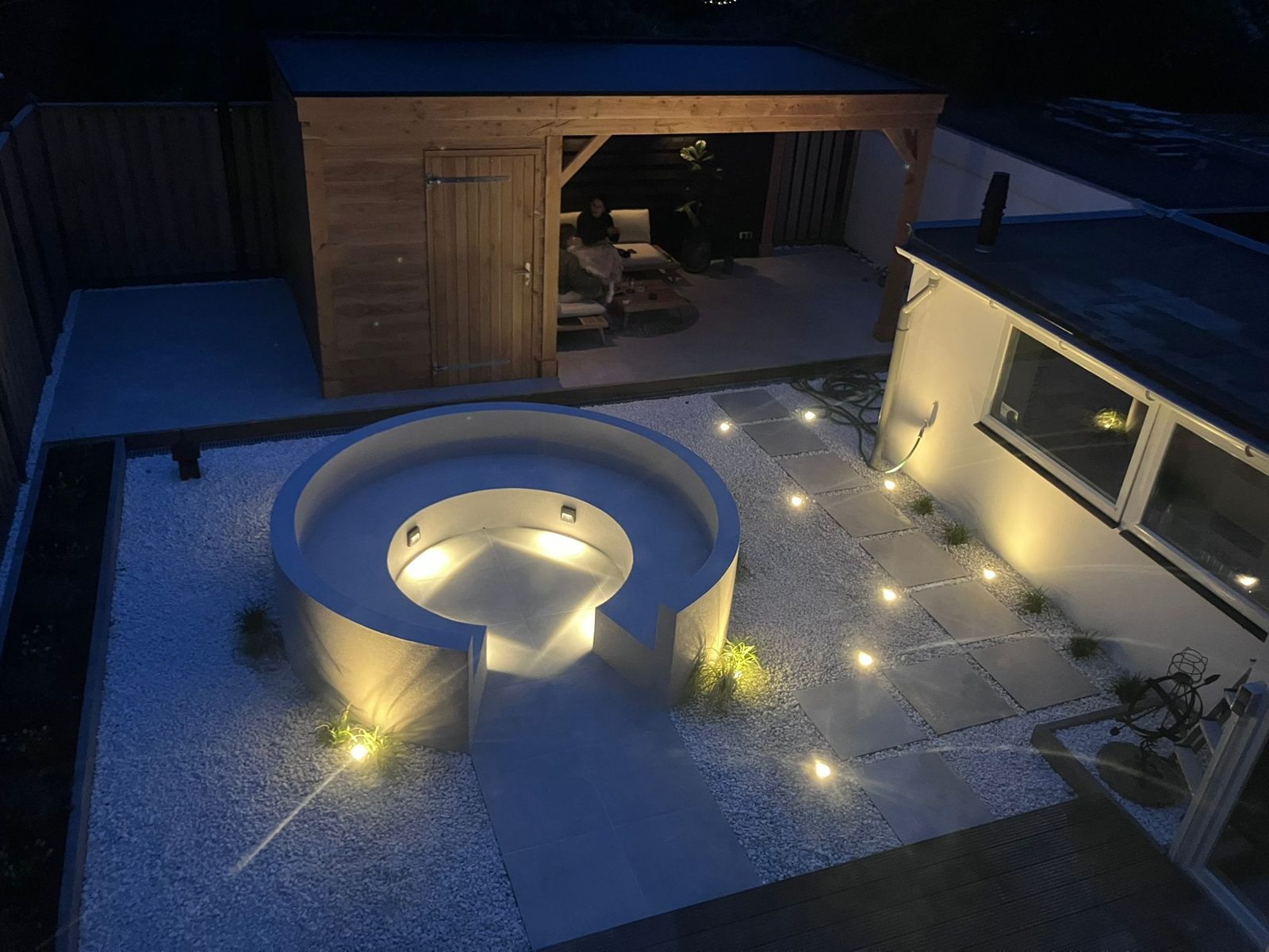 Klaar: tuin met verlichte ronde zitkuil, lichte bestrating met grondspots en een houten tuinhuis